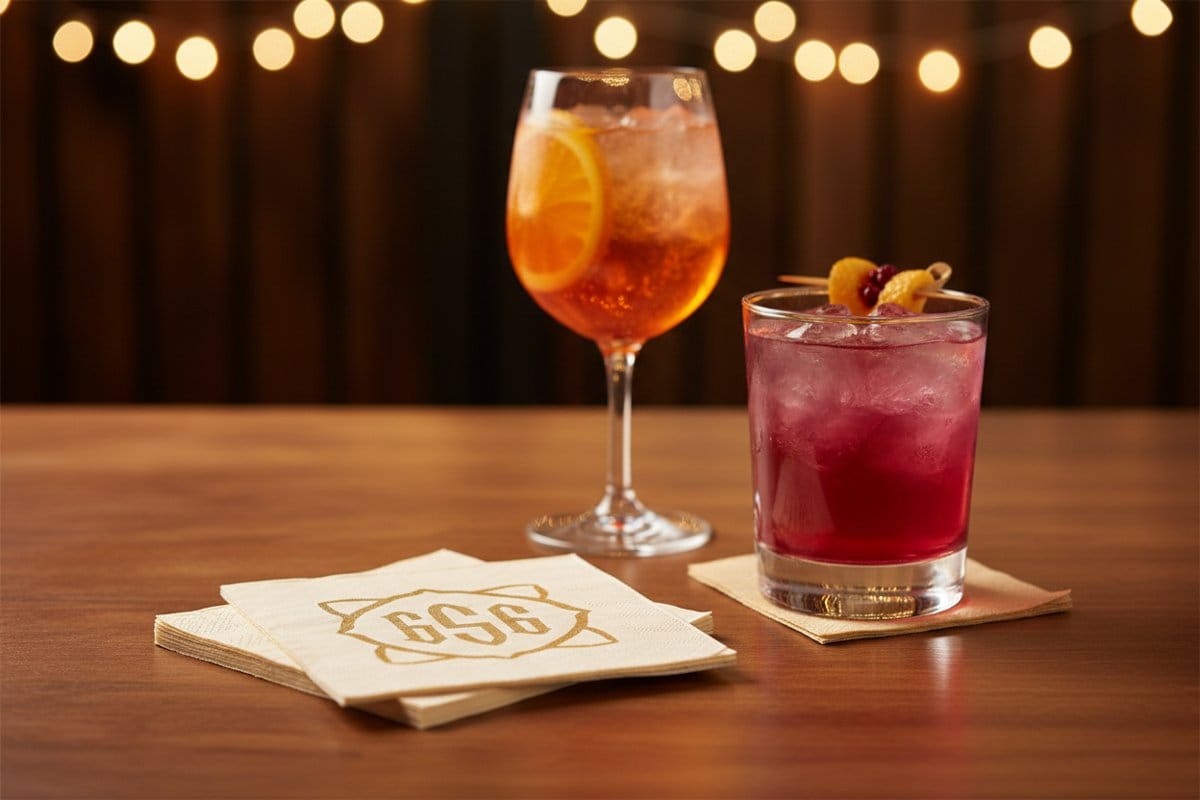 Custom printed cocktail party napkins with a gold monogram styled beside two craft cocktails on a festive bar table