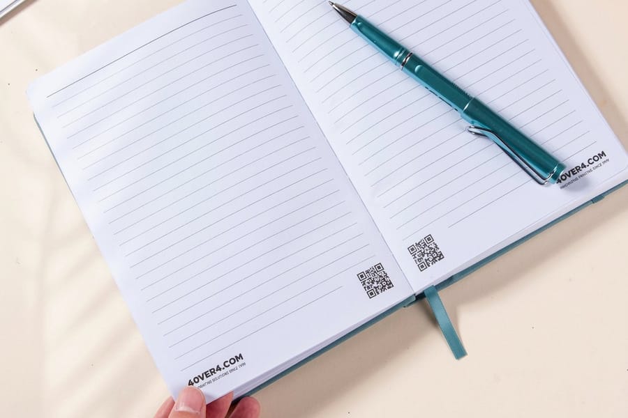 An open lined journal from 4OVER4.COM with a teal pen resting on the page and a ribbon bookmark